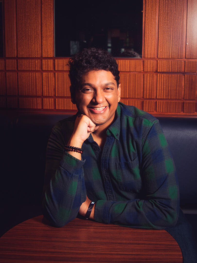 A photo of RK smiling and leaning on a table. Their fist is supporting their head and they are wearing a blue and green flannel.